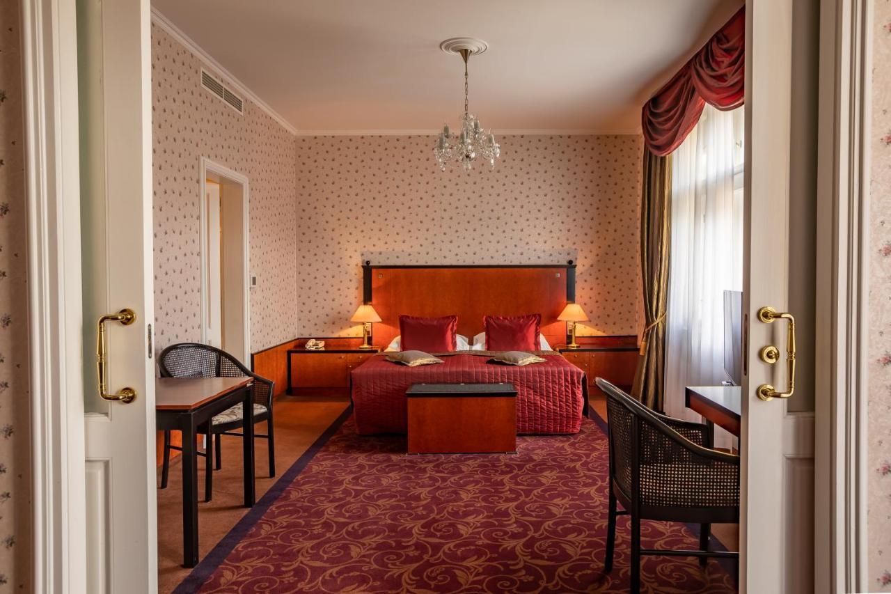 Grandhotel Pupp Karlovy Vary Bilik gambar The photo shows the interior of a hotel room. Upon entering, you can see a spacious area with a plush, red carpet. The room features a large bed with decorative pillows and a wooden headboard, flanked by bedside tables with lamps. In front of the bed