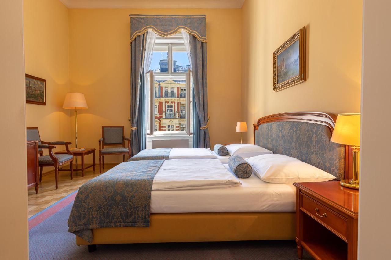 Grandhotel Pupp Karlovy Vary Luaran gambar The photo depicts a cozy hotel room or bedroom. It features a neatly made bed with a blue and white color scheme, including decorative pillows. On one side of the bed is a small bedside table with a lamp. The room has warm yellow walls and a large wi