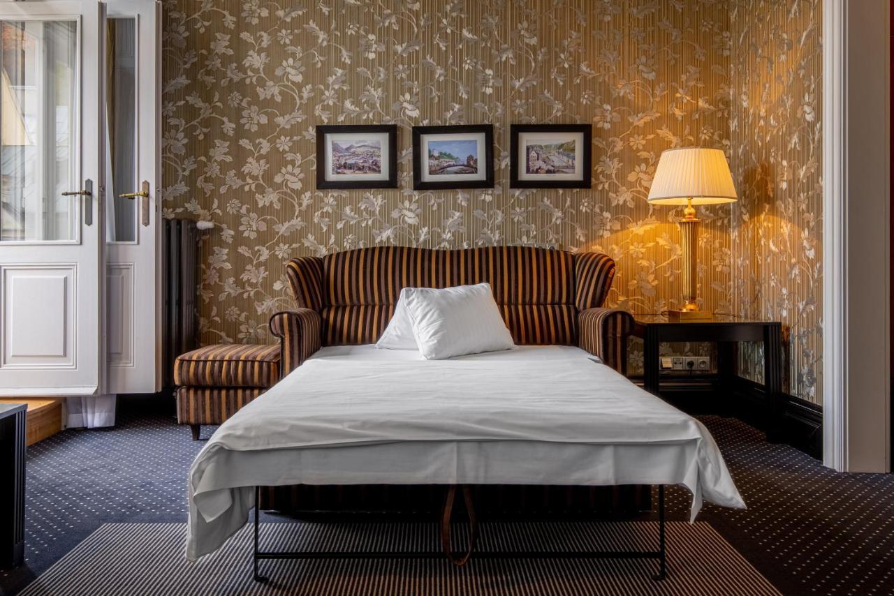 Grandhotel Pupp Karlovy Vary Luaran gambar The photo depicts a cozy room featuring a striped sofa with a white pillow on it. In front of the sofa, there is a bed or a sleeping surface with a white sheet covering it. The walls are adorned with floral wallpaper, and there are three framed pictu