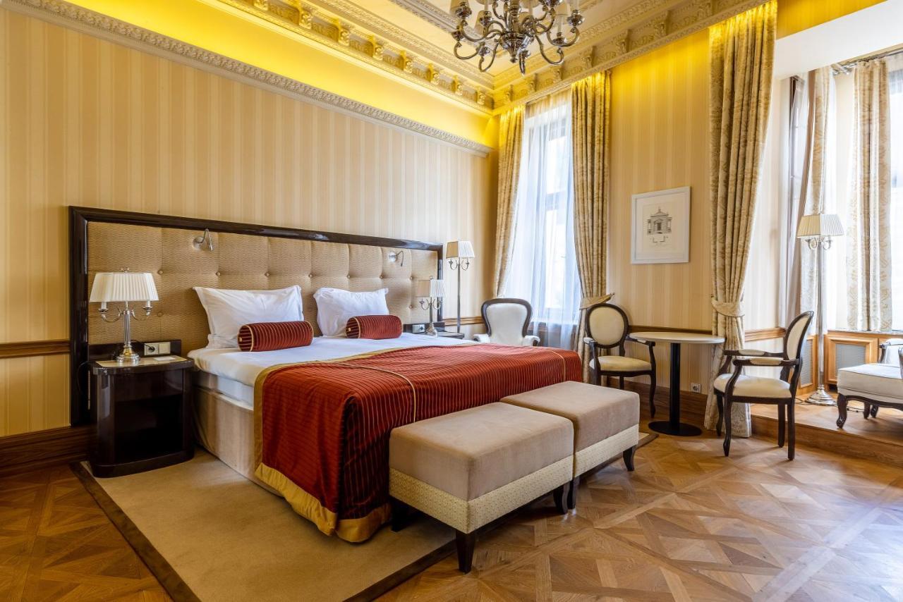 Grandhotel Pupp Karlovy Vary Luaran gambar The photo shows a luxurious hotel room designed with an elegant interior. The room features a large bed with a tufted headboard, adorned with white bedding and a deep red throw. On either side of the bed, there are bedside lamps and a small seating a