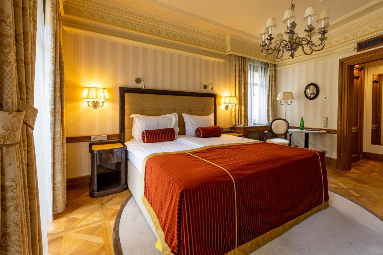 Grandhotel Pupp Karlovy Vary Luaran gambar The photo shows a hotel room featuring a large bed with an ornate red and gold bedspread, flanked by decorative pillows. The walls are subtly patterned, and there are two lamps mounted on either side of the bed. A chandelier hangs from the ceiling, a
