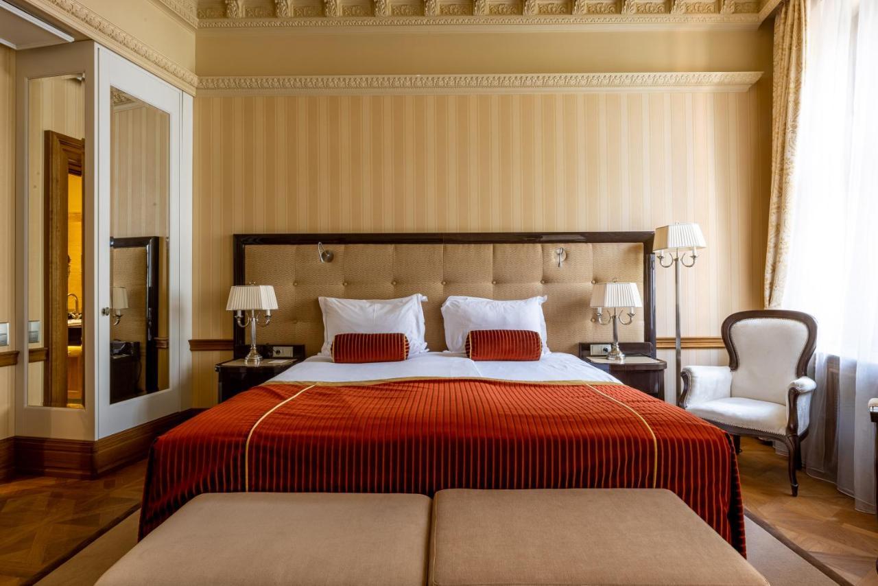 Grandhotel Pupp Karlovy Vary Luaran gambar The photo shows an elegantly decorated hotel room featuring a large bed with a plush headboard. The bed is dressed in rich, orange bedding, complemented by white pillows and decorative cushions. Flanking the bed are two bedside lamps on matching nigh