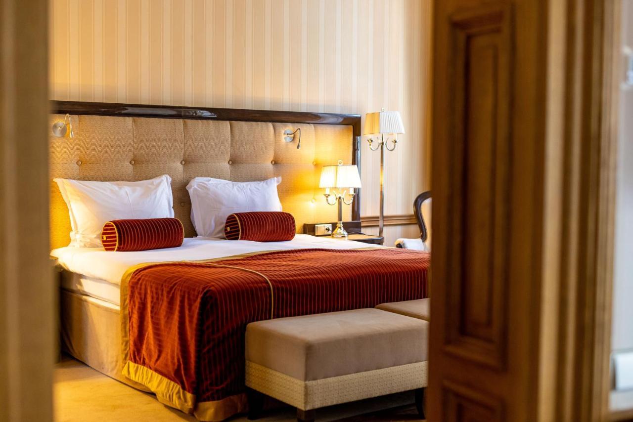 Grandhotel Pupp Karlovy Vary Luaran gambar The photo shows a well-furnished hotel room with a cozy and elegant design. It features a large bed with white bedding and decorative orange-red throw pillows at the headboard. The walls are light-colored with subtle stripes, adding a touch of sophis