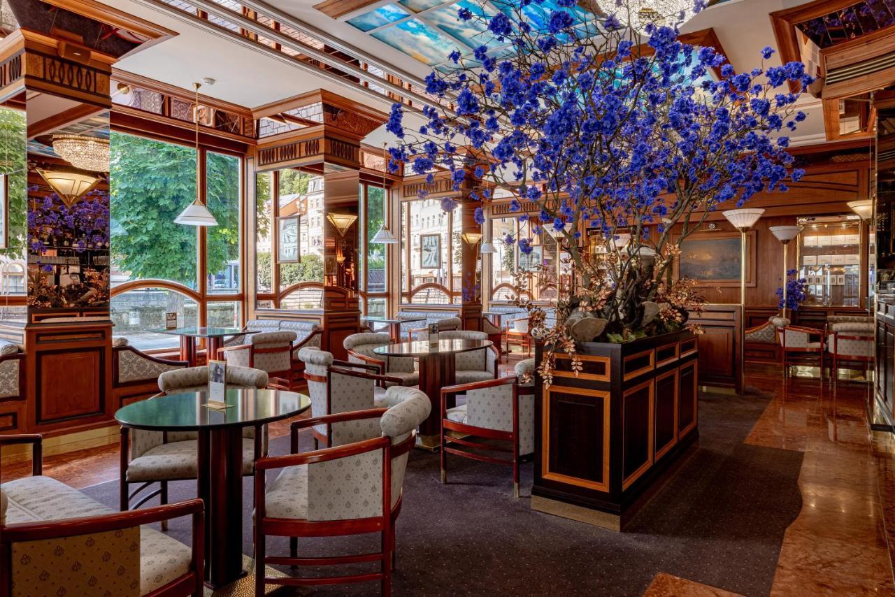 Grandhotel Pupp Karlovy Vary Luaran gambar The photo shows an elegant interior of a restaurant or café. It features wooden furnishings with intricate details, including circular tables and upholstered chairs. A large floral arrangement, predominantly featuring blue flowers, is placed in the c