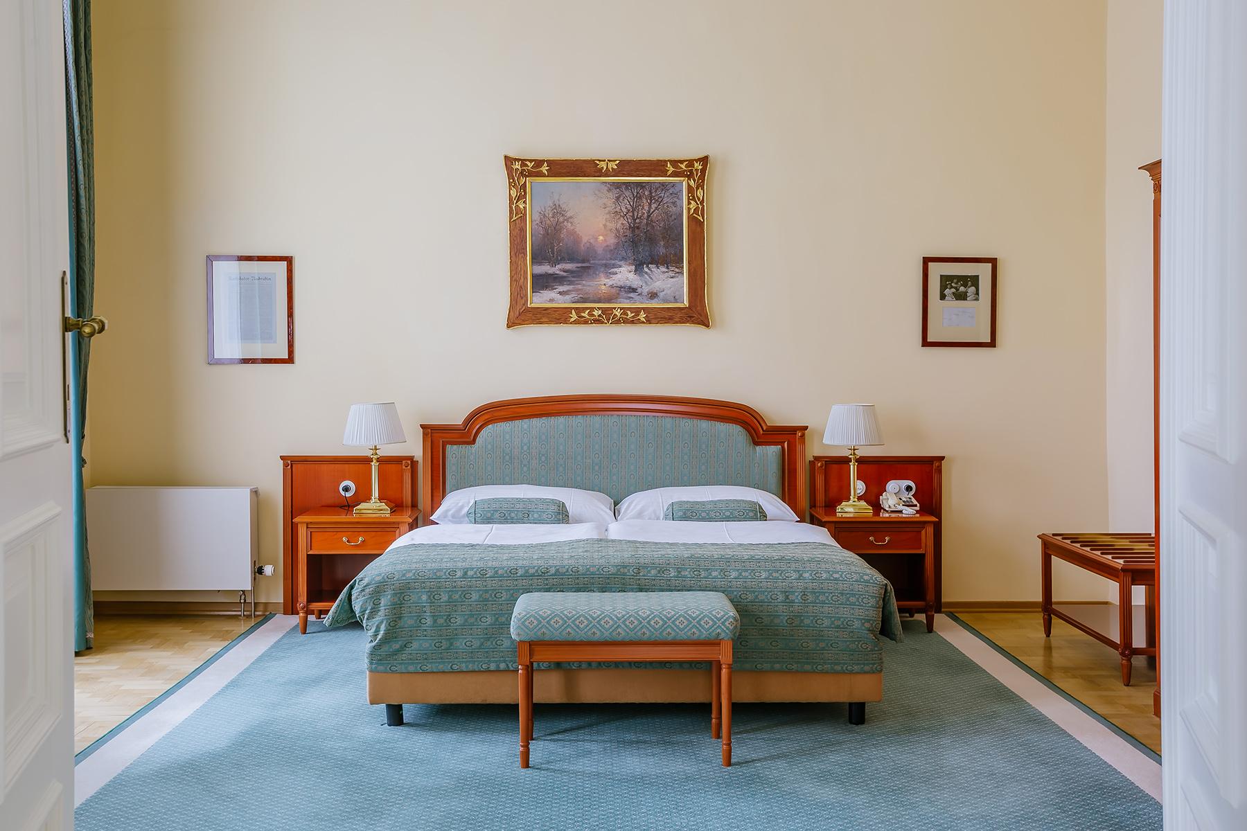 Grandhotel Pupp Karlovy Vary Luaran gambar The photo shows a cozy bedroom with a light-colored wall. There is a large bed with a muted green coverlet and white bedding. On either side of the bed, there are bedside tables with lamps. At the foot of the bed, there is a small bench. Above the be