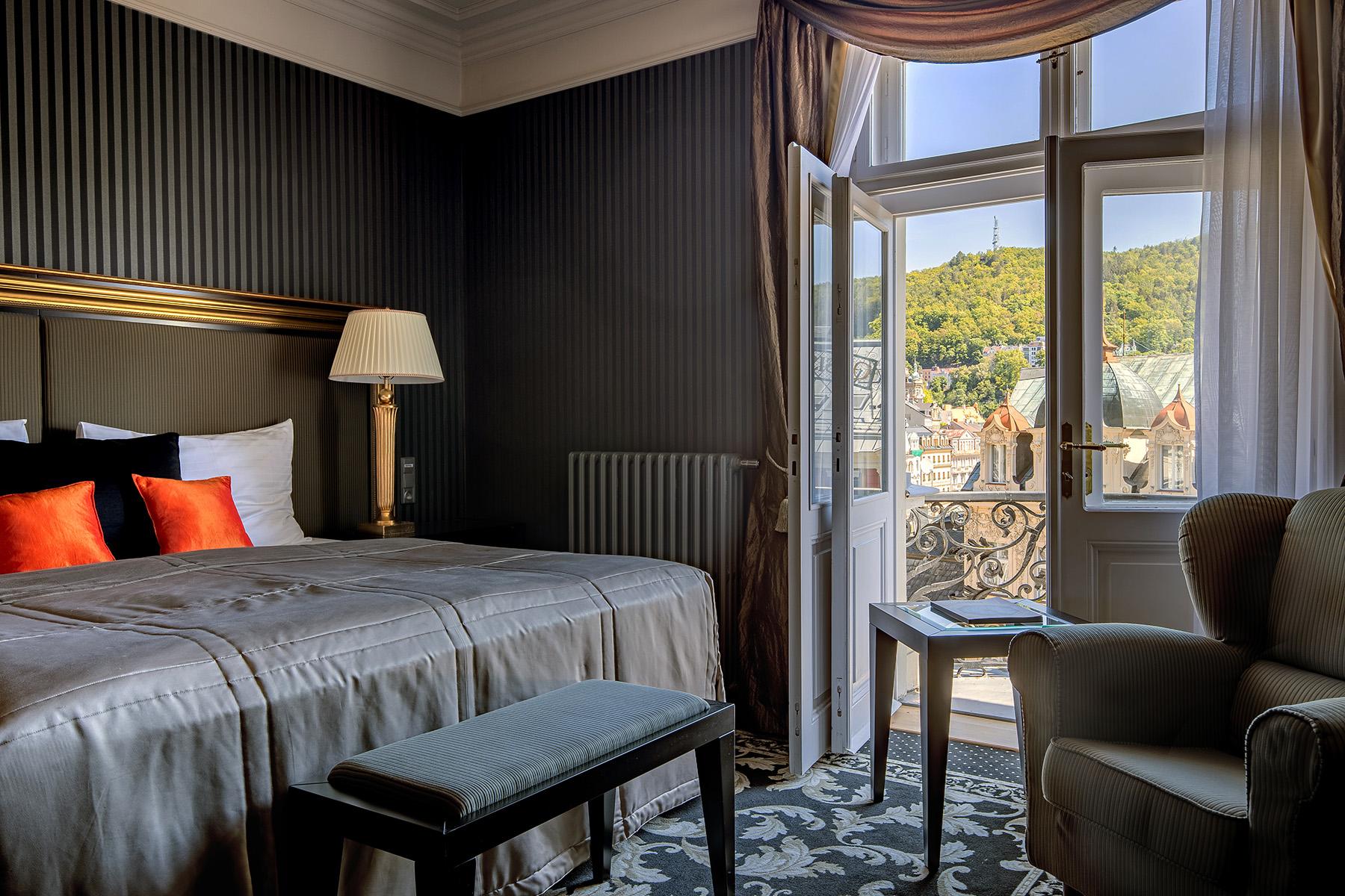 Grandhotel Pupp Karlovy Vary Luaran gambar The photo shows a cozy hotel room with a sophisticated design. The room features a bed with a plush grey duvet and decorative pillows in black and orange. There is a bedside lamp beside the bed. A comfortable armchair sits in one corner. The room has
