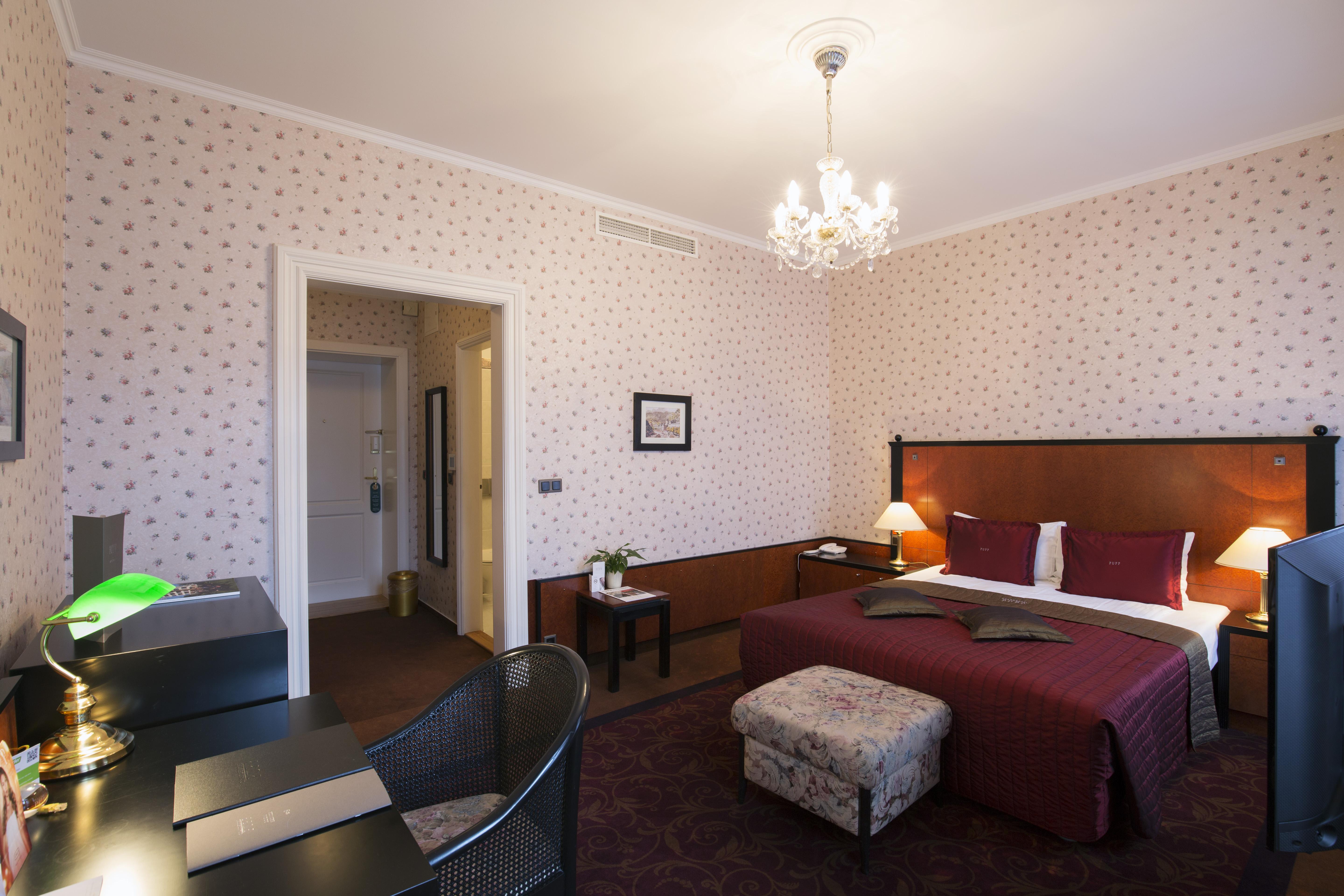 Grandhotel Pupp Karlovy Vary Luaran gambar The photo depicts a well-furnished hotel room. There is a large bed with a burgundy bedspread, complemented by decorative pillows. The room features soft lighting from a chandelier and lamps on either side of the bed. 

To the left, there is a desk w