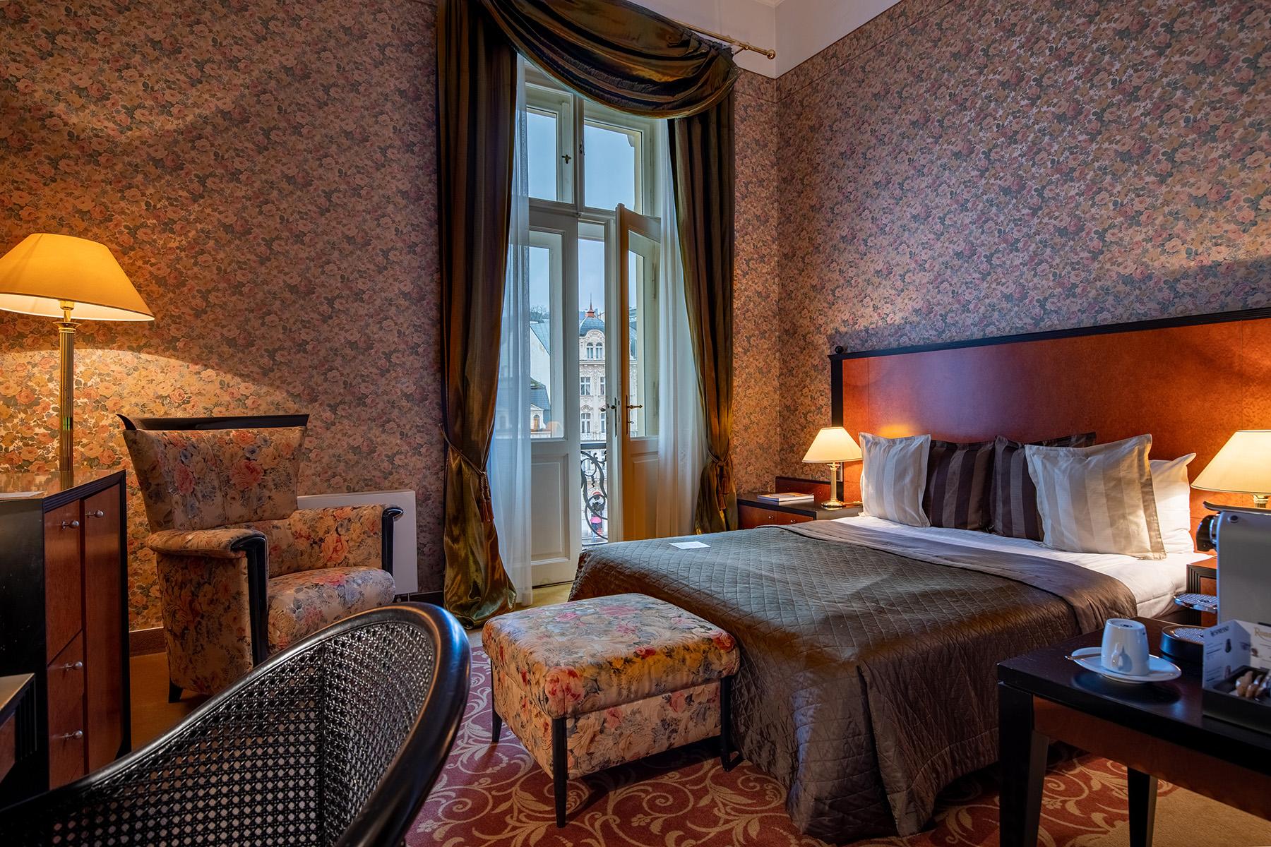 Grandhotel Pupp Karlovy Vary Luaran gambar The photo shows a cozy hotel room featuring a queen-sized bed with dark bedding and several decorative pillows. There is a comfortable armchair and an ottoman in a floral pattern, adding a touch of elegance. A wooden bedside table holds a lamp and a 