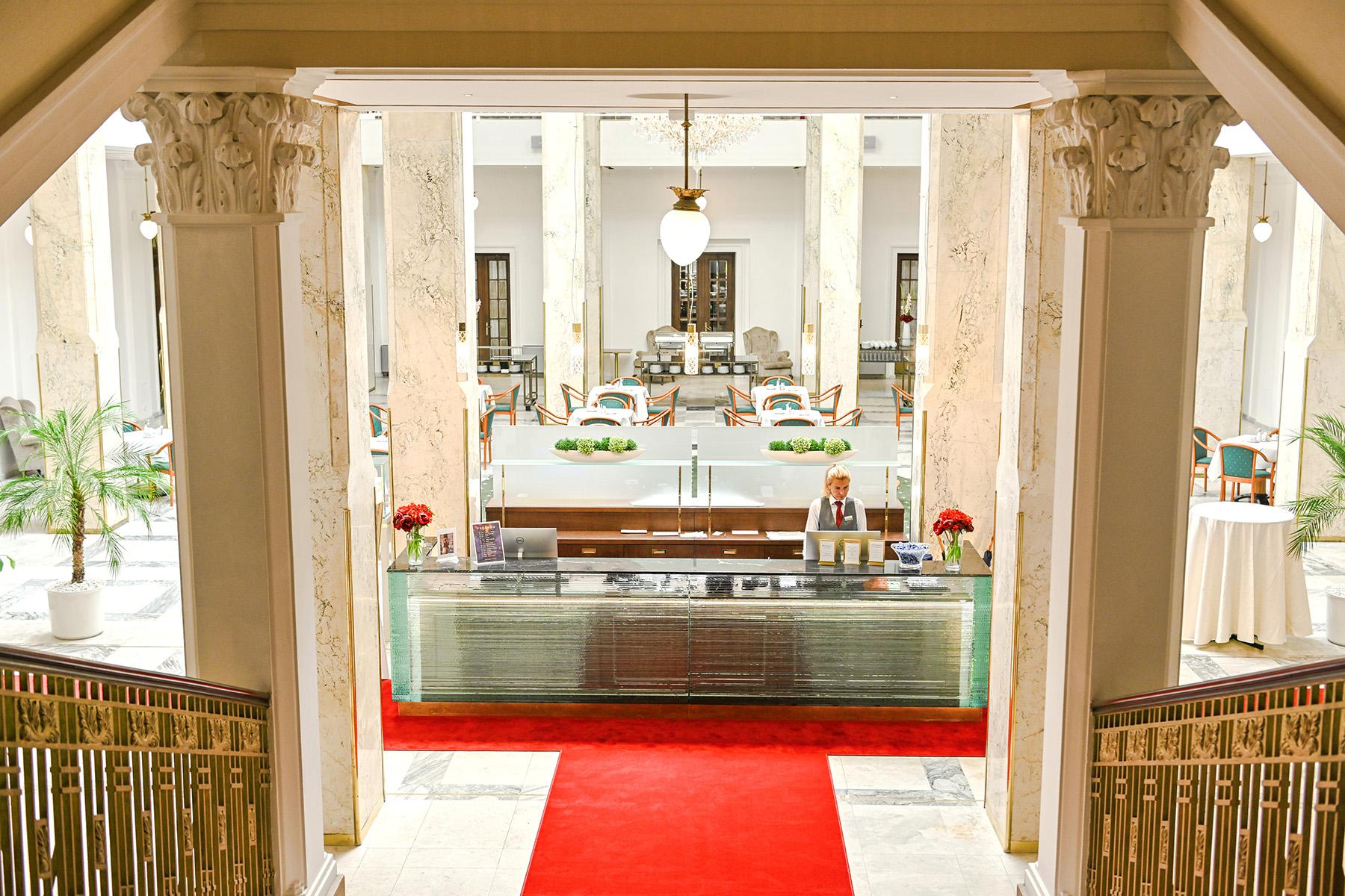 Grandhotel Pupp Karlovy Vary Luaran gambar The photo shows the interior of a hotel lobby. There is a reception desk made of glass and wood, situated at the front, with a staff member standing behind it. The lobby features elegant columns and a high ceiling, giving it a grand appearance. A red