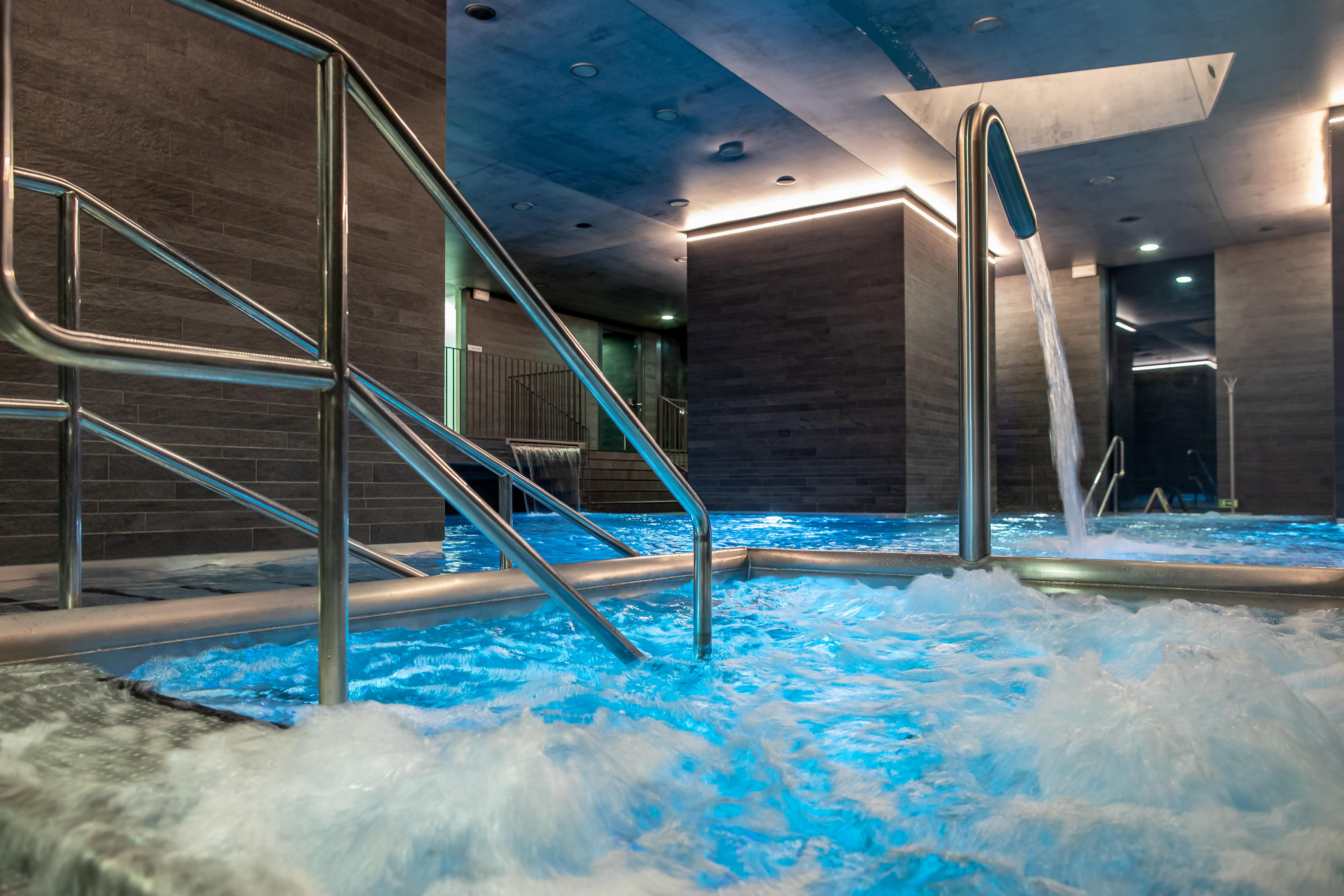 Grandhotel Pupp Karlovy Vary Luaran gambar The photo shows a sleek, modern spa area featuring a hot tub or pool with bubbling, illuminated blue water. There are stainless steel handrails leading into the water. The walls have a contemporary design with a gray finish, and there are soft lights