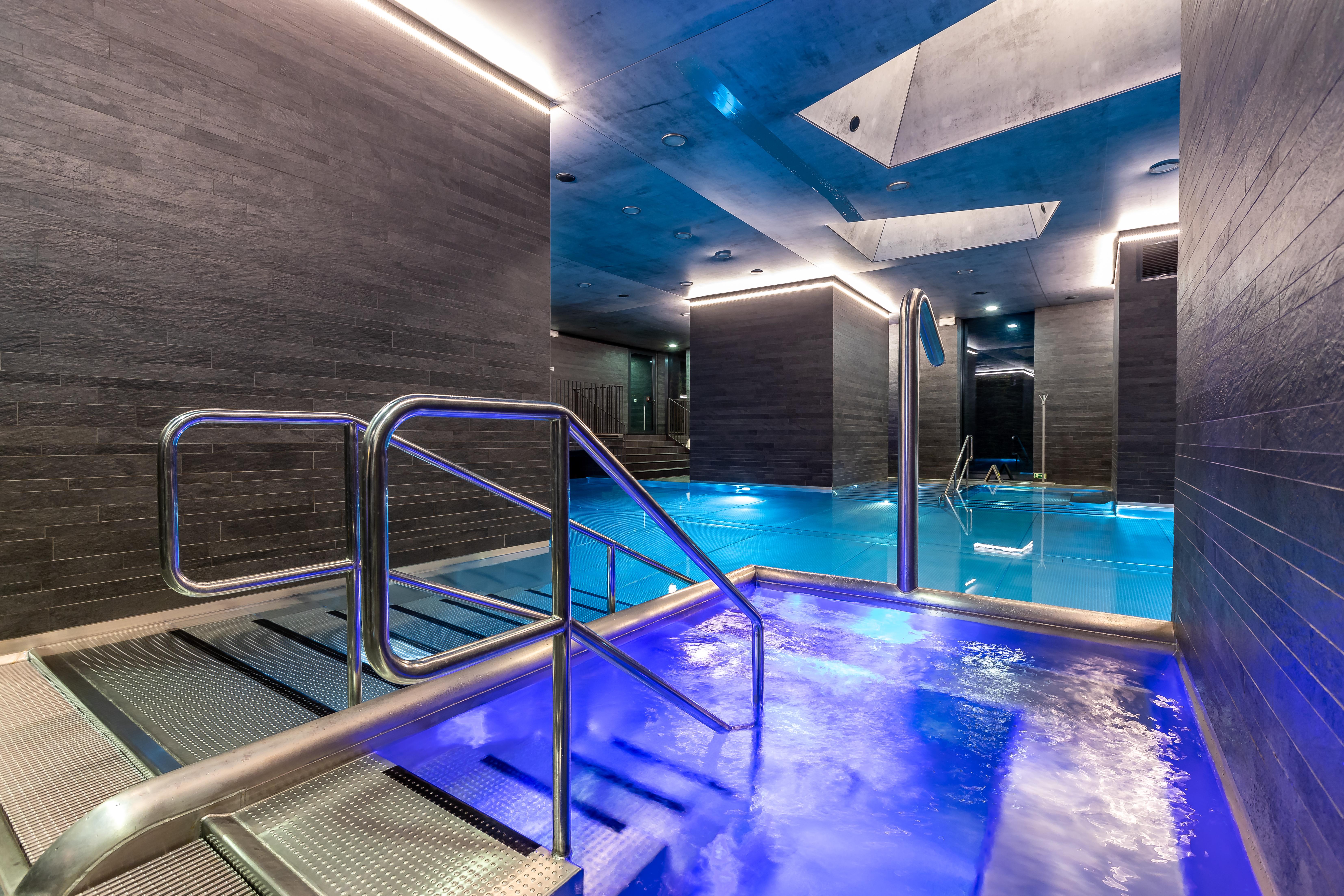 Grandhotel Pupp Karlovy Vary Luaran gambar The photo shows a modern spa area with a pool. There are sleek, dark walls and blue lighting that creates a tranquil atmosphere. A metal railing leads down into the pool, which has a section with bubbling water. The overall design features clean line