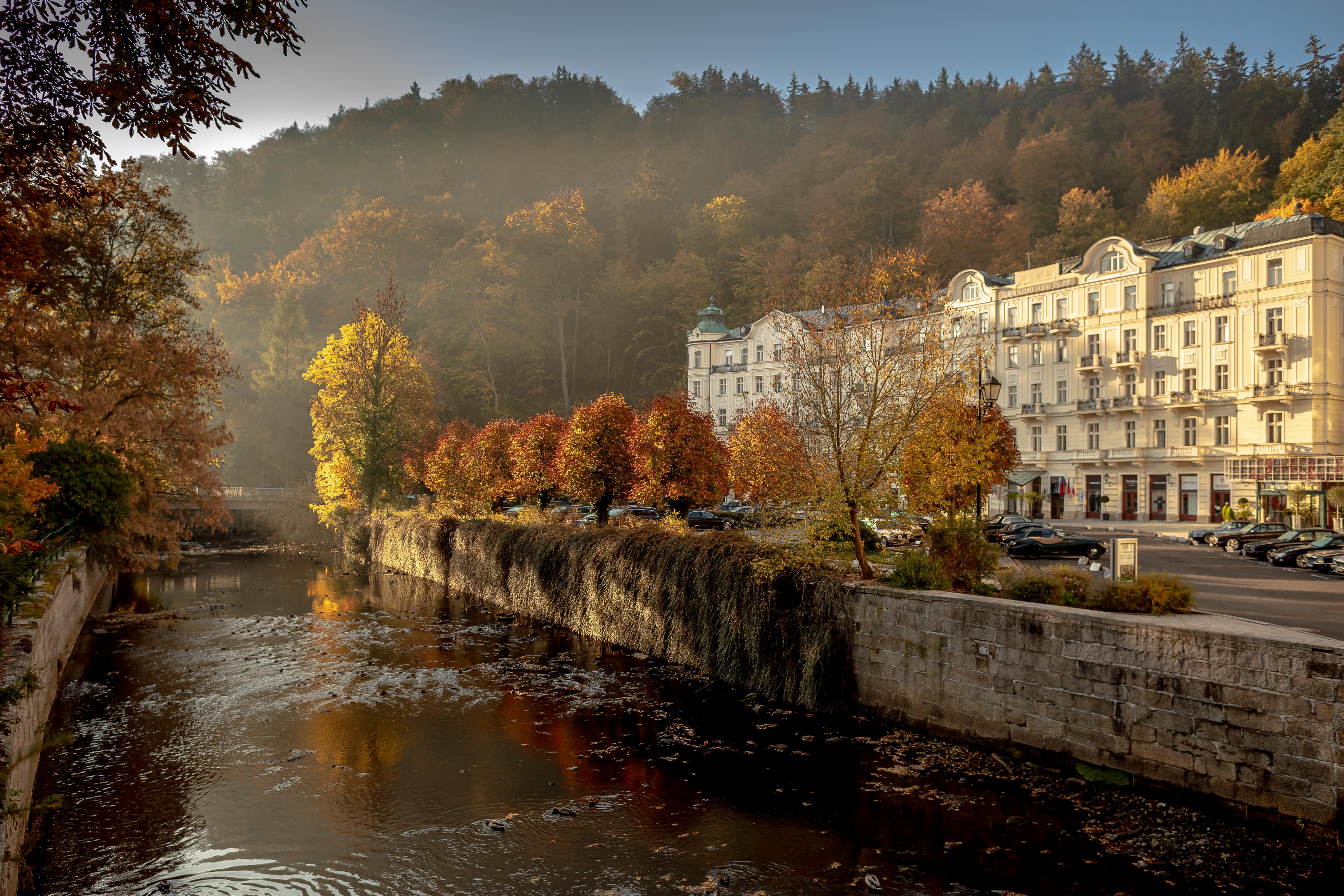 Grandhotel Pupp Karlovy Vary Luaran gambar The photo depicts a serene riverside scene during autumn. The river flows gently, reflecting the warm hues of the trees along its banks. On one side, there are vibrant orange and yellow trees, showcasing the beautiful fall foliage, while the opposite