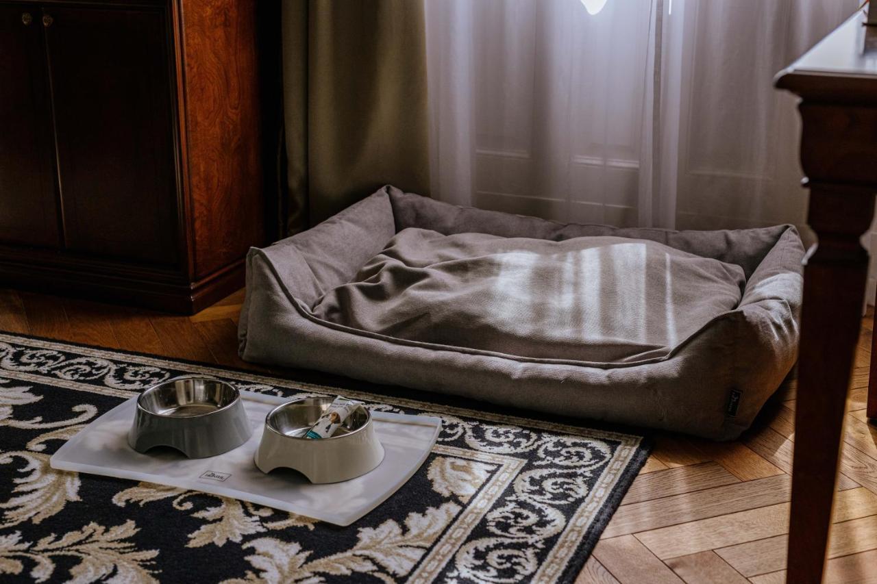 Grandhotel Pupp Karlovy Vary Luaran gambar The photo shows a cozy pet area inside a room. There is a soft, rectangular pet bed in a muted color, positioned against a wall, with some light filtering through a window. In front of the bed, there are two pet bowls placed on a mat, one bowl is gre