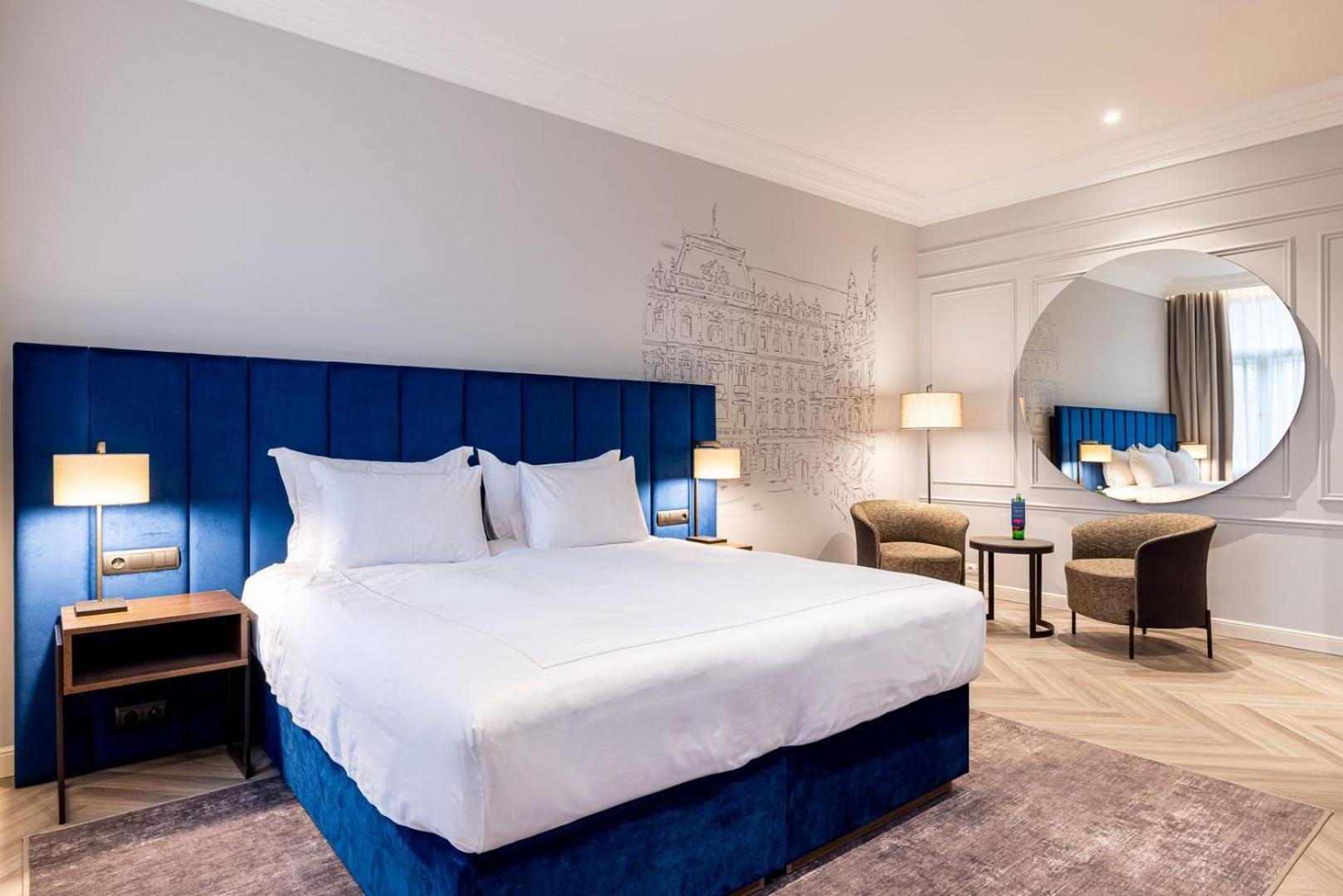 Grandhotel Pupp Karlovy Vary Luaran gambar The photo shows a modern hotel room featuring a large double bed with white linens and a blue upholstered headboard. The walls are light gray, and there is a decorative mural in the background. Two bedside lamps are on either side of the bed. In the 