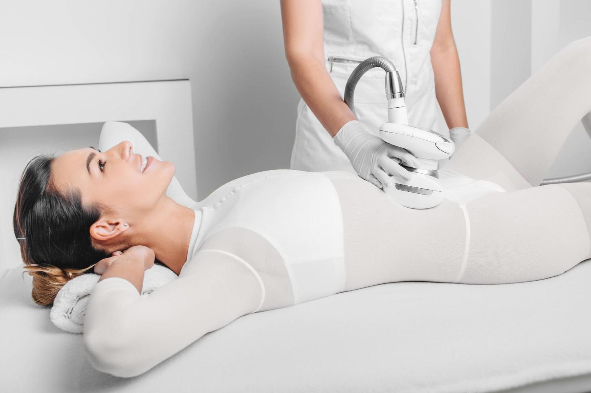 Grandhotel Pupp Karlovy Vary Luaran gambar The photo depicts a woman lying on a treatment table, appearing relaxed and smiling. She is wearing a form-fitting white outfit, likely designed for a cosmetic or wellness procedure. A technician, dressed in a white outfit and gloves, is applying a d