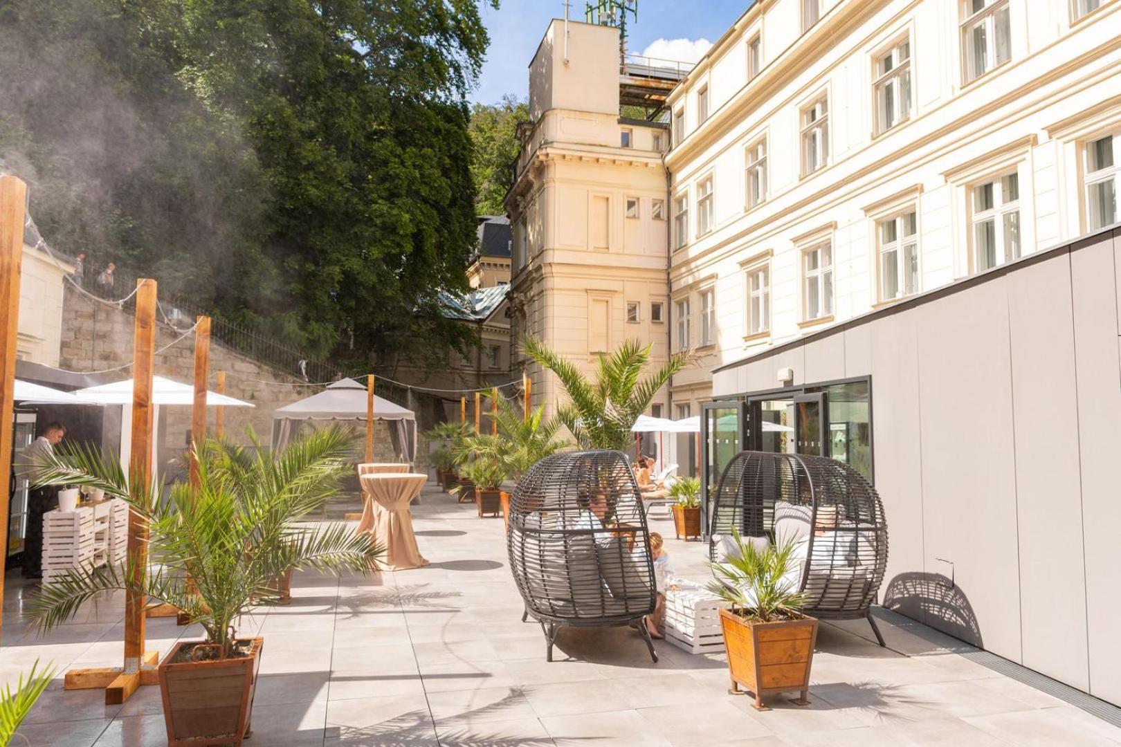 Grandhotel Pupp Karlovy Vary Luaran gambar The photo shows a stylish outdoor area with a mix of modern and classic architecture. There are several large potted plants, likely palm trees, which add a tropical feel to the space. Two round, open wicker chairs are placed on a paved surface, provi