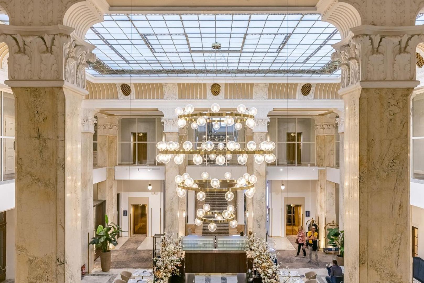 Grandhotel Pupp Karlovy Vary Luaran gambar The photo depicts an elegant hotel lobby characterized by its spacious and luxurious design. Large marble columns rise to support a ceiling with a glass skylight, allowing natural light to illuminate the area. A central chandelier made of numerous ro
